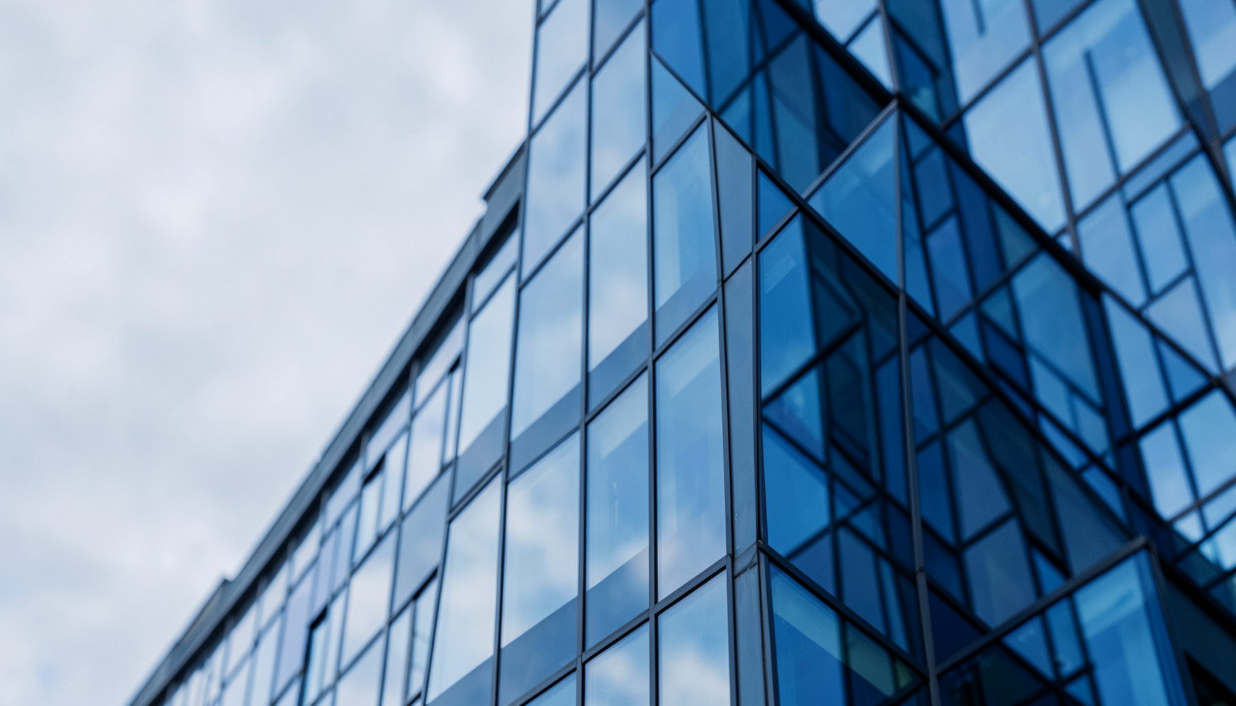 Modern bir ofis binasının cam cephesi, açık gri gökyüzüne karşı yansımalar; kurumsal ve güvenilir bir görünüm.